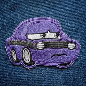 Puede incluir: Un parche bordado p&uacute;rpura de un coche de dibujos animados con una expresi&oacute;n triste. El coche tiene faros blancos, una parrilla negra y detalles en negro. El parche est&aacute; sobre un fondo de mezclilla azul.