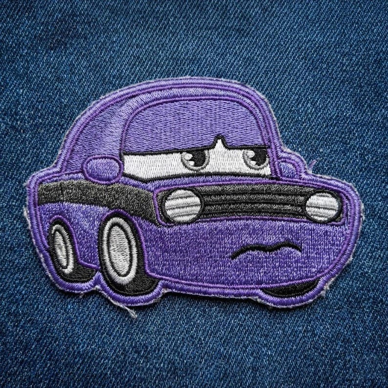 Puede incluir: Un parche bordado p&uacute;rpura de un coche de dibujos animados con una expresi&oacute;n triste. El coche tiene faros blancos, una parrilla negra y detalles en negro. El parche est&aacute; sobre un fondo de mezclilla azul.