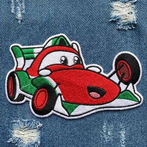 Puede incluir: Parche bordado de un coche de carreras de dibujos animados en rojo, blanco y verde. El coche tiene ojos grandes y amigables y una boca sonriente. El parche est&aacute; sobre un fondo de mezclilla azul.