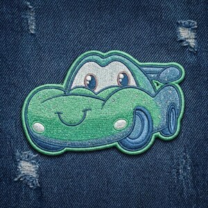 Puede incluir: Parche bordado de coche verde y azul con una cara sonriente, faros blancos y ruedas azules. El parche est&aacute; sobre un fondo de mezclilla azul.