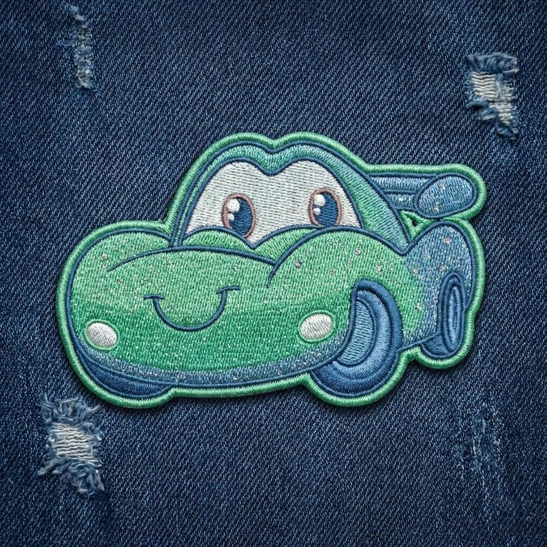 Puede incluir: Parche bordado de coche verde y azul con una cara sonriente, faros blancos y ruedas azules. El parche est&aacute; sobre un fondo de mezclilla azul.