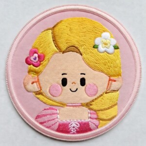 May include: Embroidered patch depicting a cartoon girl with long, flowing blonde hair. The girl has rosy cheeks, a smiling face, and wears a pink dress. The patch is round with a pink border and floral accents.