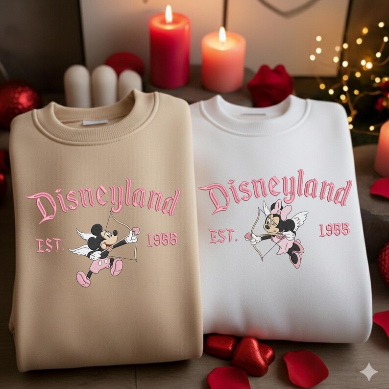 May include: Two sweatshirts, one tan and one white, featuring the word "Disneyland" in pink script. Each sweatshirt has a graphic of Mickey and Minnie Mouse as cupids, with the year "1955" below. The sweatshirts are part of a Disney-themed collection.
