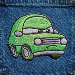 Puede incluir: Un parche de coche bordado verde con un dise&ntilde;o de dibujos animados, con una cara sonriente y ruedas grandes. El parche est&aacute; cosido sobre una tela vaquera azul, mostrando los detalles de los faros, la parrilla y las ventanas del coche.
