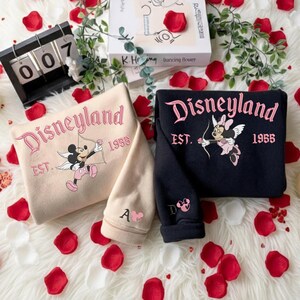 May include: Two sweatshirts, one beige and one navy blue, with the word "Disneyland" in pink script. The beige sweatshirt features Mickey Mouse as Cupid, and the navy blue sweatshirt features Minnie Mouse as Cupid. Both sweatshirts have "EST. 1955" printed below the Disneyland text.