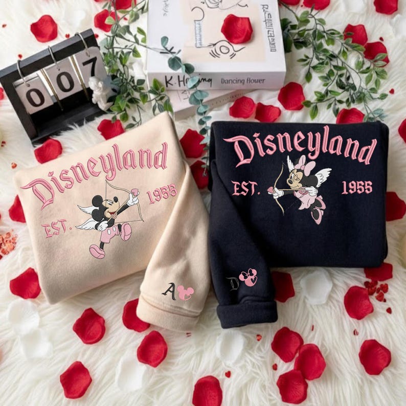 May include: Two sweatshirts, one beige and one navy blue, with the word "Disneyland" in pink script. The beige sweatshirt features Mickey Mouse as Cupid, and the navy blue sweatshirt features Minnie Mouse as Cupid. Both sweatshirts have "EST. 1955" printed below the Disneyland text.