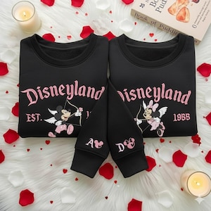 May include: Two black sweatshirts with the word "Disneyland" in pink script, featuring Mickey and Minnie Mouse as cupids. The sleeves have initials and a heart. The sweatshirts are surrounded by rose petals and candles.