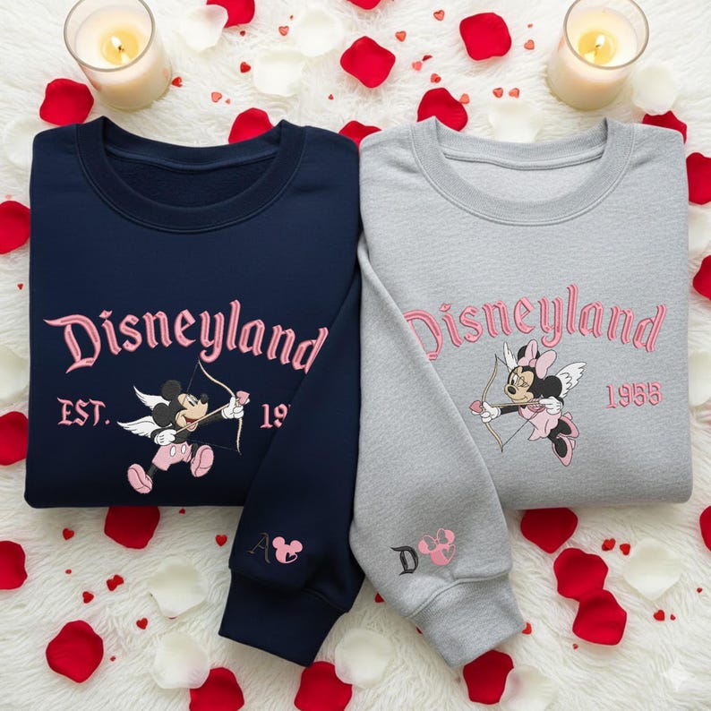 May include: Two sweatshirts, one navy blue and one gray, with the word "Disneyland" in pink script. Each sweatshirt features a cartoon character with a bow and arrow. The navy sweatshirt has "EST. 19" and the gray one has "1955".