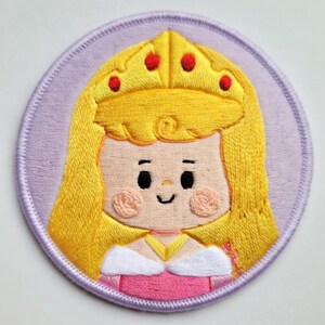 May include: Embroidered patch of a cartoon princess with long yellow hair, a golden crown with red jewels, and a pink dress. The patch is circular, with a light purple background and a stitched border.
