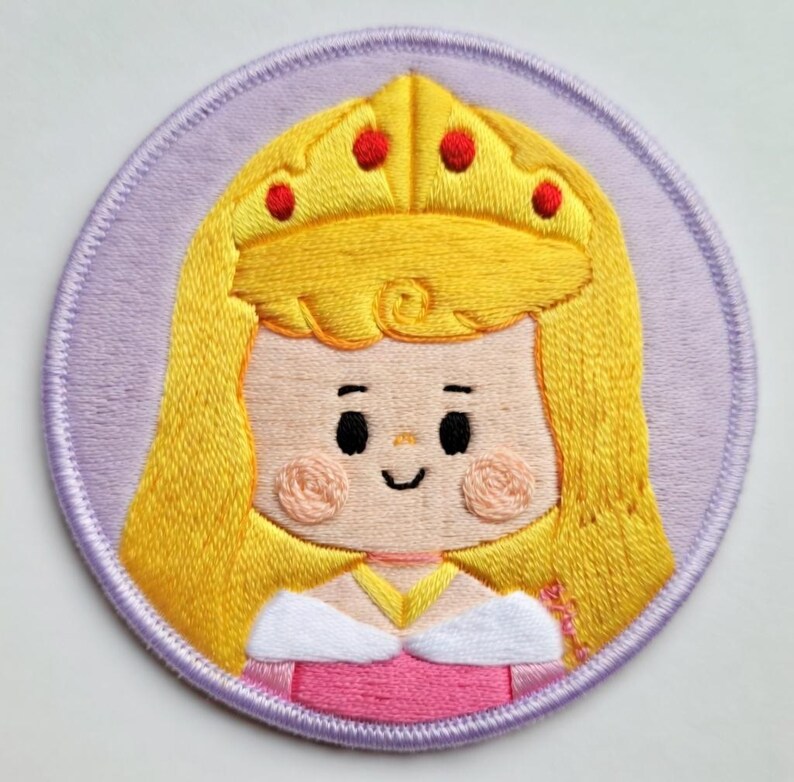 May include: Embroidered patch of a cartoon princess with long yellow hair, a golden crown with red jewels, and a pink dress. The patch is circular, with a light purple background and a stitched border.