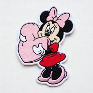 Op de afbeelding: Geborduurde patch met Minnie Mouse in een rode jurk en roze strik, met een roze hart. De patch heeft een witte rand. Een kleurrijk ontwerp voor kleding of accessoires.