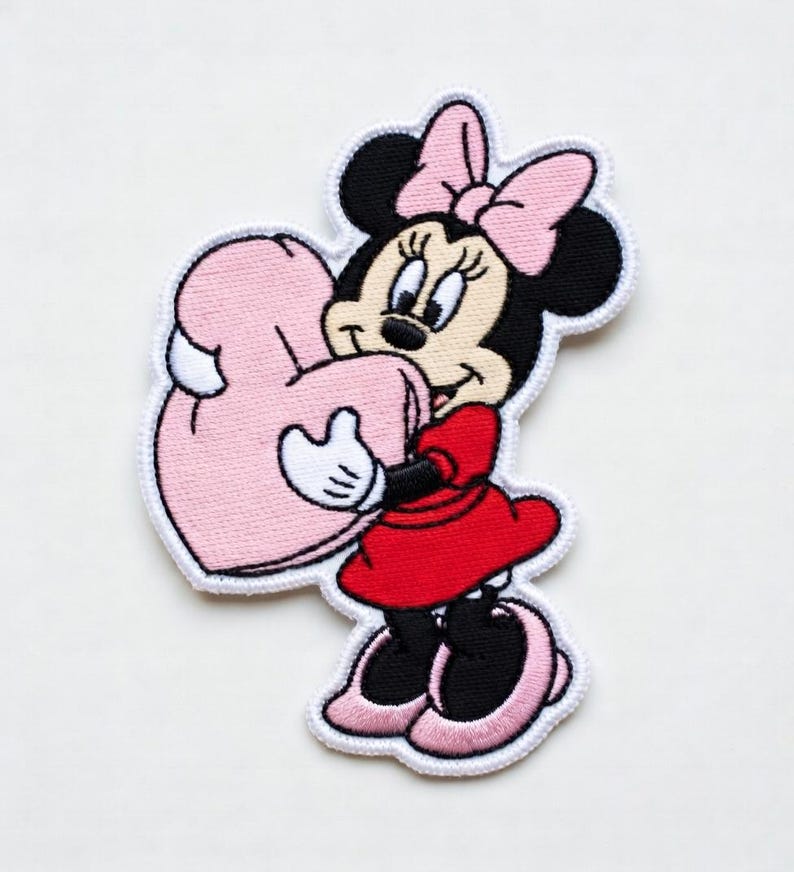 Op de afbeelding: Geborduurde patch met Minnie Mouse in een rode jurk en roze strik, met een roze hart. De patch heeft een witte rand. Een kleurrijk ontwerp voor kleding of accessoires.