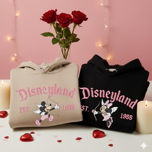 May include: Two hooded sweatshirts, one tan and one black, with the word "Disneyland" embroidered in pink script. Each sweatshirt features a cartoon character with a bow and arrow. Red roses and candles are in the background.
