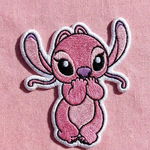 Puede incluir: Parche bordado con un personaje de dibujos animados rosa con grandes ojos negros, una nariz morada y detalles blancos. El personaje est&aacute; sentado, con las manos cubriendo su boca, sobre un fondo de tela rosa.