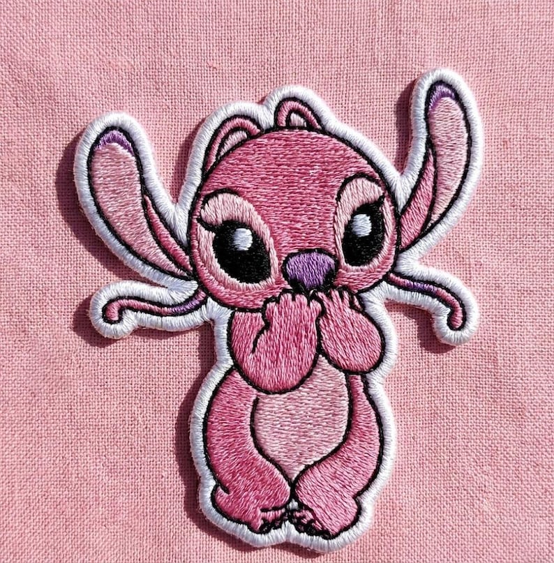 Puede incluir: Parche bordado con un personaje de dibujos animados rosa con grandes ojos negros, una nariz morada y detalles blancos. El personaje est&aacute; sentado, con las manos cubriendo su boca, sobre un fondo de tela rosa.