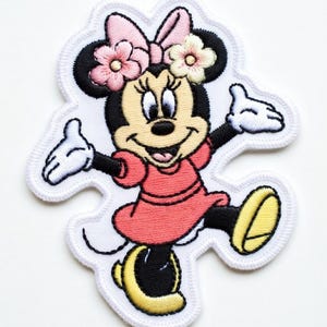 Op de afbeelding: Geborduurde patch met Minnie Mouse in een vrolijke pose. Minnie draagt een rode jurk, gele schoenen en een roze strik met bloemen. De patch heeft een witte rand.