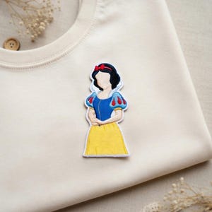 May include: Embroidered patch featuring a cartoon illustration of Snow White in a blue top with red and white sleeves, a yellow skirt, and a red bow. The patch is on a cream-colored fabric.