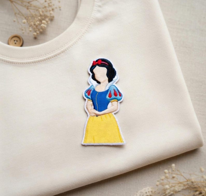 May include: Embroidered patch featuring a cartoon illustration of Snow White in a blue top with red and white sleeves, a yellow skirt, and a red bow. The patch is on a cream-colored fabric.