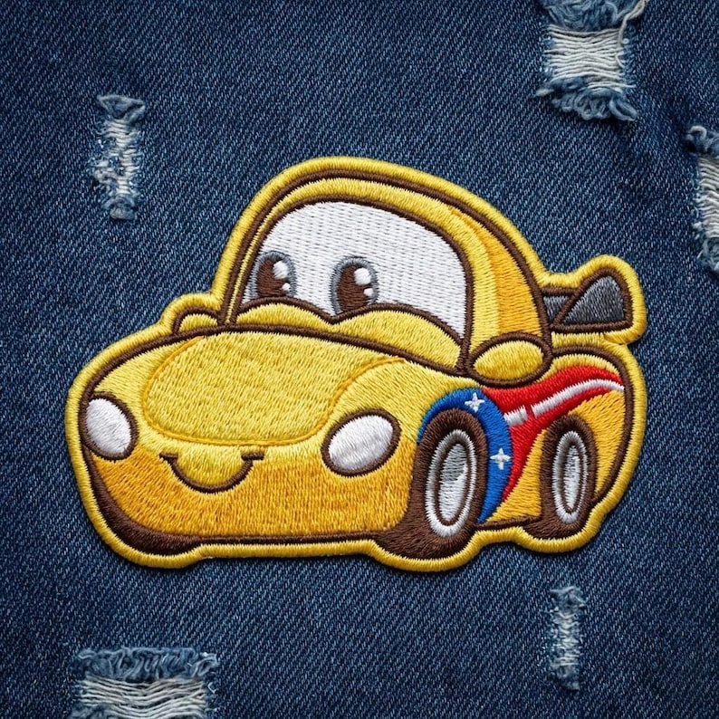 Puede incluir: Parche bordado de un coche de dibujos animados amarillo sonriente con neum&aacute;ticos marrones y un dise&ntilde;o rojo y azul en el lateral. El parche est&aacute; sobre un fondo de mezclilla azul desgastado.