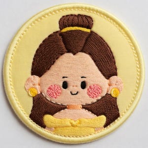 May include: Embroidered patch depicting a cartoon character with brown hair, pink cheeks, and a yellow dress. The round patch has a yellow border and a stitched design.