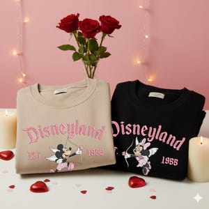 May include: Two sweatshirts, one tan and one black, with the word "Disneyland" in pink script. Each sweatshirt features a cartoon character with a bow and arrow. The tan sweatshirt has Mickey Mouse, and the black one has Minnie Mouse. The year "1955" is also embroidered.