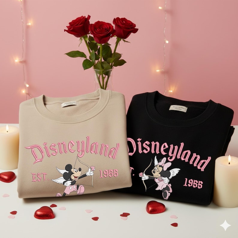May include: Two sweatshirts, one tan and one black, with the word "Disneyland" in pink script. Each sweatshirt features a cartoon character with a bow and arrow. The tan sweatshirt has Mickey Mouse, and the black one has Minnie Mouse. The year "1955" is also embroidered.