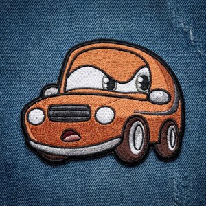 Puede incluir: Parche de coche naranja bordado con un dise&ntilde;o de dibujos animados. El coche tiene ojos grandes, una parrilla negra y ruedas marrones. El parche est&aacute; delineado en negro y sobre un fondo de mezclilla azul.