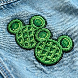 May include: Two green embroidered patches in the shape of Mickey Mouse's head, with a waffle design. The patches are set against a light blue denim background, with a black border and circular ear details.