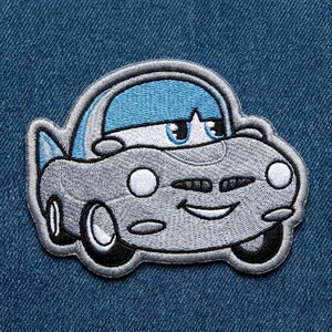 Puede incluir: Un parche bordado con un coche de dibujos animados con una cara sonriente. El coche es principalmente gris con un techo azul y detalles blancos. El parche est&aacute; sobre un fondo de mezclilla azul.