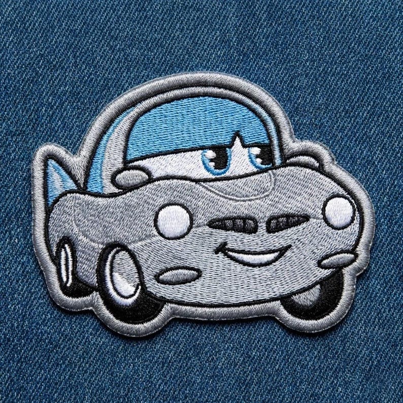 Puede incluir: Un parche bordado con un coche de dibujos animados con una cara sonriente. El coche es principalmente gris con un techo azul y detalles blancos. El parche est&aacute; sobre un fondo de mezclilla azul.