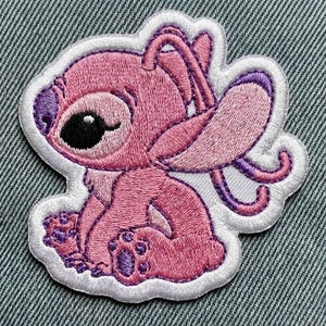 Puede incluir: Parche bordado con un personaje de dibujos animados rosa con orejas grandes, nariz morada y un ojo negro. El parche tiene un borde blanco y probablemente sea para ropa o accesorios. El personaje est&aacute; sentado con las piernas cruzadas.