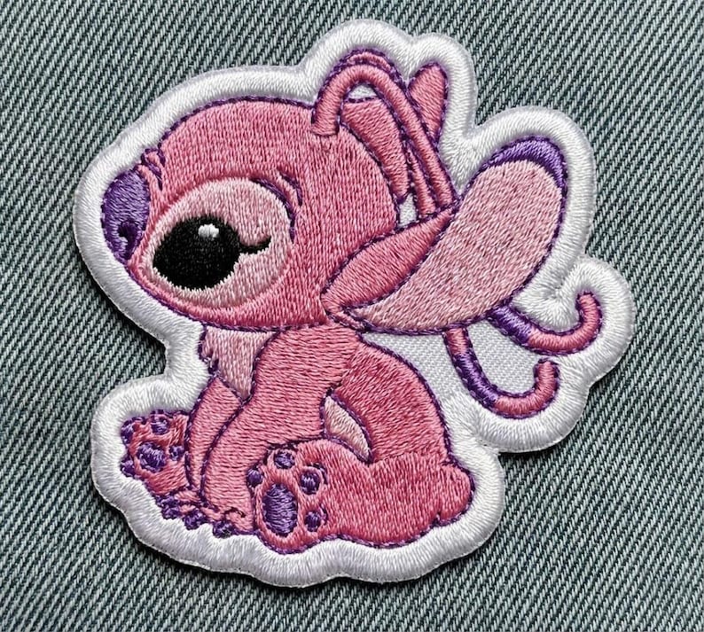 Puede incluir: Parche bordado con un personaje de dibujos animados rosa con orejas grandes, nariz morada y un ojo negro. El parche tiene un borde blanco y probablemente sea para ropa o accesorios. El personaje est&aacute; sentado con las piernas cruzadas.