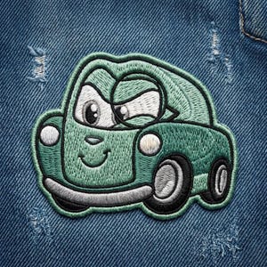 Puede incluir: Parche bordado de un coche de dibujos animados verde sonriente, con ojos grandes. El parche tiene contornos negros y est&aacute; sobre un fondo de mezclilla azul. El coche tiene dos faros y dos ruedas.