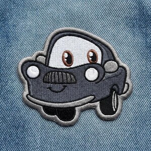 Puede incluir: Parche bordado de un coche de dibujos animados con una cara sonriente. El coche es principalmente gris oscuro con detalles blancos y negros, incluyendo las ruedas, los faros y un parabrisas blanco. El parche est&aacute; sobre un fondo de mezclilla azul.