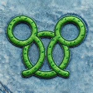 Puede incluir: Un parche bordado verde con un diseño de ocho repetido. El parche está cosido con hilo verde brillante y presenta un aspecto texturizado. El parche está sobre un fondo de mezclilla azul claro.