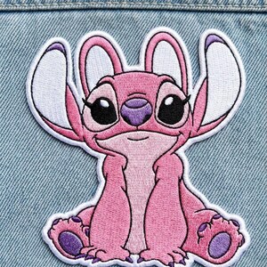 Puede incluir: Parche bordado rosa con un personaje de dibujos animados con grandes ojos negros, nariz morada y orejas con puntas blancas. El personaje est&aacute; sentado con los brazos y las piernas extendidos. El parche est&aacute; sobre un fondo de mezclilla.