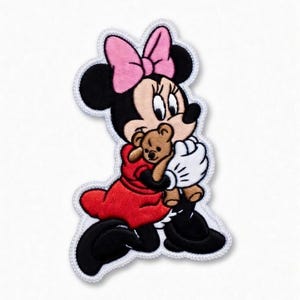 Op de afbeelding: Geborduurde patch met een stripfiguur met een roze strik, zwarte oren en een rode jurk, die een kleine bruine teddybeer vasthoudt. Het personage heeft witte handschoenen en zwarte schoenen, met een witte rand.