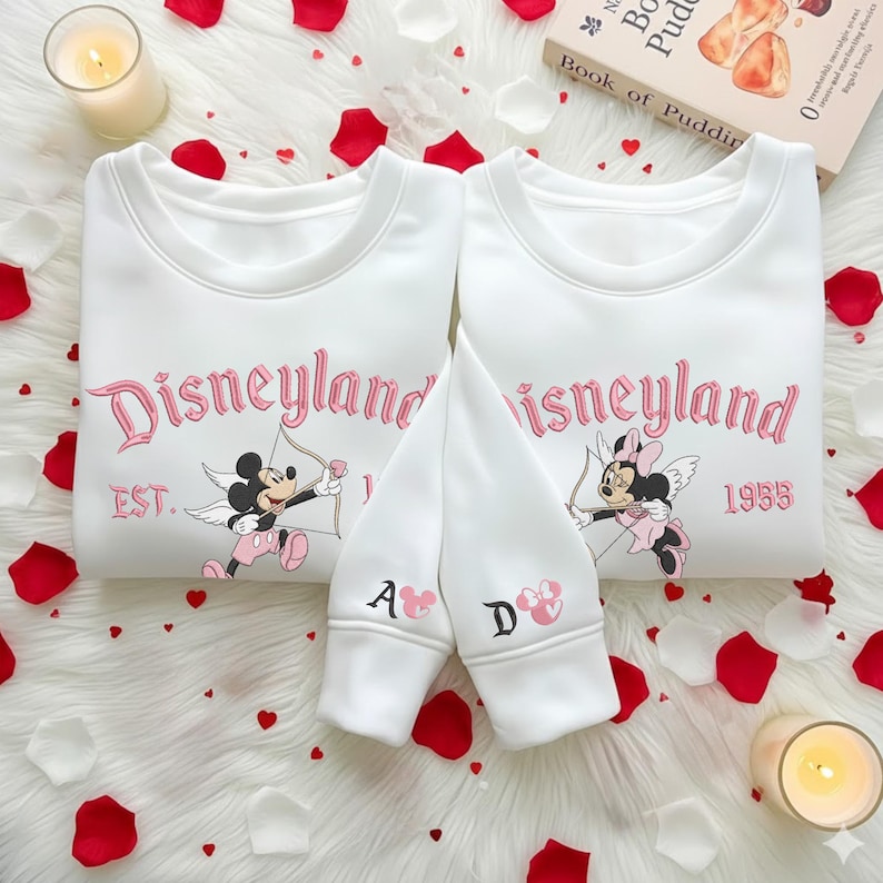 May include: Two white sweatshirts with the word "Disneyland" in pink script, featuring Mickey and Minnie Mouse as cupids. The sleeves have the letters "A" and "D" with heart accents. The sweatshirts are surrounded by rose petals and candles.