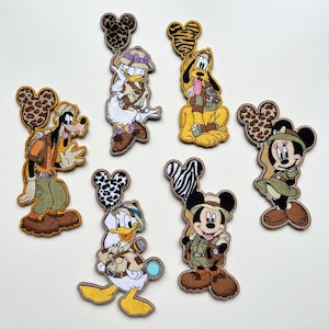 Può includere: Toppe ricamate raffiguranti personaggi Disney in tenute da safari. Pippo, Paperino, Pluto, Topolino e Minnie sono raffigurati in varie pose, con cappelli e attrezzature da safari. Ogni toppa ha un palloncino a forma di testa di Topolino.