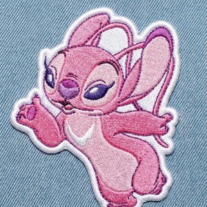 Puede incluir: Parche bordado rosa con el personaje Angel de la pel&iacute;cula Lilo & Stitch. El parche tiene un borde blanco y est&aacute; sobre un fondo de mezclilla azul claro. Angel se representa con orejas grandes, ojos morados y una pose acogedora.