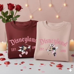 May include: Two sweatshirts, one burgundy and one blush pink, featuring the Disneyland logo in pink script. Each sweatshirt has a cartoon character with a bow and arrow. The burgundy sweatshirt has Mickey Mouse, and the pink one has Minnie Mouse.