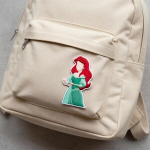 May include: Beige canvas backpack with a front pocket and a zipper closure. A colorful embroidered patch of a cartoon character with red hair and a green dress is attached to the front pocket.