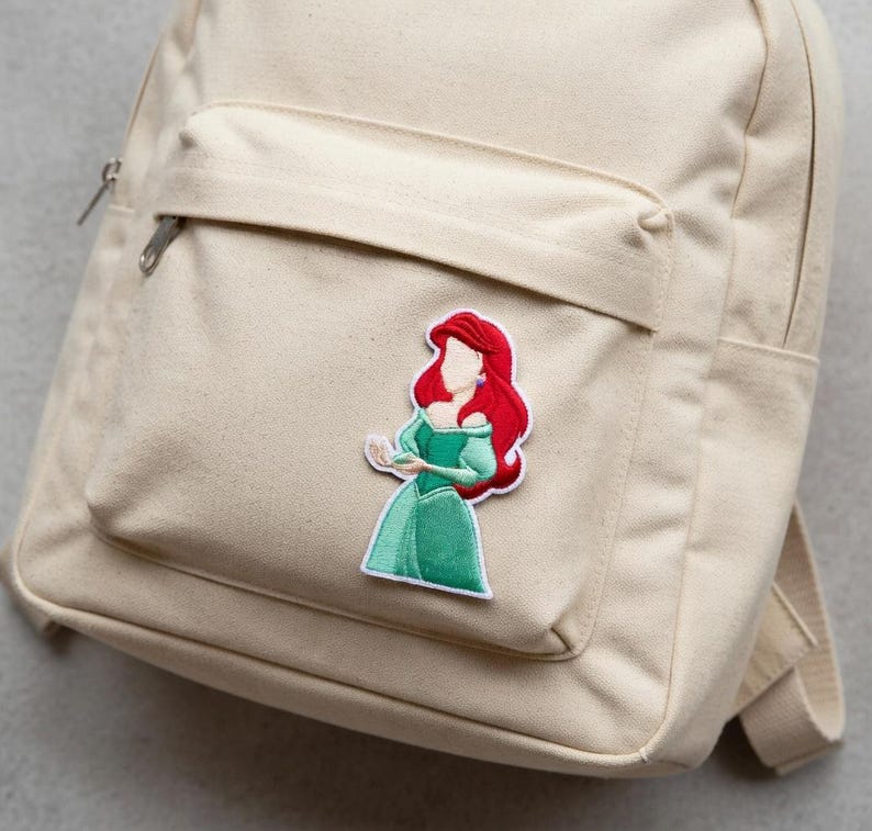 May include: Beige canvas backpack with a front pocket and a zipper closure. A colorful embroidered patch of a cartoon character with red hair and a green dress is attached to the front pocket.
