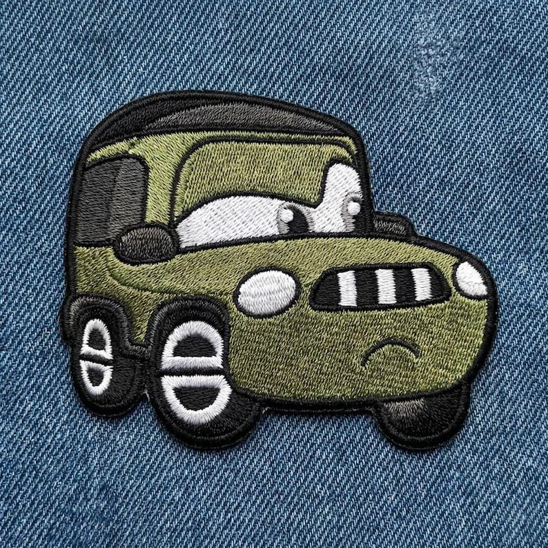 Puede incluir: Un parche bordado con un coche de dibujos animados en verde oliva, negro y blanco. El coche tiene ojos grandes y una boca sonriente. El parche est&aacute; dise&ntilde;ado para planchar o coser sobre ropa o telas.