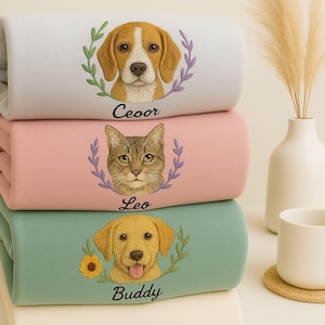 Pode incluir: Uma pilha de três cobertores bordados em cores pastel. O cobertor superior é cinza claro com um desenho de beagle e o nome "Ceoor". O cobertor do meio é rosa com um desenho de gato e o nome "Leo". O cobertor inferior é verde-azulado com um desenho de golden retriever e o nome "Buddy".