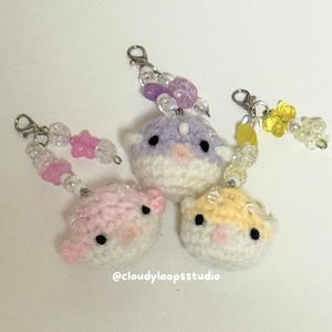 May include: Three handmade crochet hamster keychains in pink, purple, and yellow. Each keychain features a small, round hamster with black eyes and a white belly. They are attached to beaded chains with silver clasps. The text "@cloudyloopsstudio" is visible.