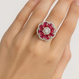 May include: A flower-shaped ring featuring a cluster of oval-cut, vibrant red gemstones surrounded by a halo of smaller, clear stones. The ring's setting appears to be silver or white gold, creating a striking contrast with the red gems.