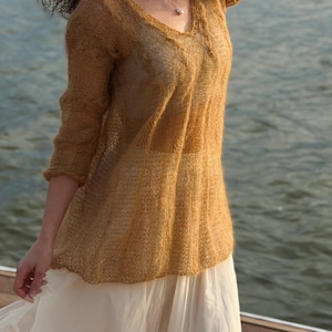 May include: A golden-brown, sheer knit top with a V-neck and three-quarter sleeves. The top is paired with a cream-colored skirt. The outfit is worn outdoors near water.