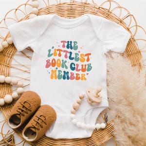 Może przedstawiać: Białe body dla dziecka z napisem "THE LITTLEST BOOK CLUB MEMBER" w kolorowej czcionce retro. Obraz zawiera brązowe buciki dziecięce, smoczek i klips do smoczka z koralikami, wszystko na tle plecionego wiklinowego kosza.