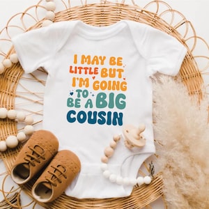 以下が含まれることがあります： オレンジ、青、緑で「I MAY BE LITTLE BUT I'M GOING TO BE A BIG COUSIN」と書かれた白いベビーボディスーツ。茶色のベビーシューズとおしゃぶりも画像にあります。アイテムは、織られた籐のマットの上に配置されています。
